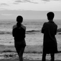 Black and white photograph of two young people standing next to each other, silhouetted against the beach and sky