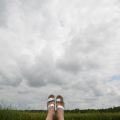 A big sky full of fluffy clouds surrounds a pair of feet in silver sandals at the bottom center of the image.