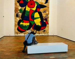 A man and woman embrace while sitting on a bench in a gallery, looking onto a large woven piece of art in the style of Jean Miro